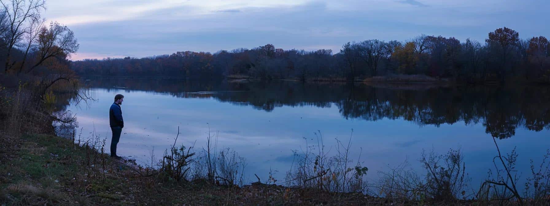 a contemplative man stands at the edge of a serene lake at dusk, reflecting on the emotional weight of stress, surrounded by soft, diffused twilight hues that evoke a sense of introspection and tranquility.