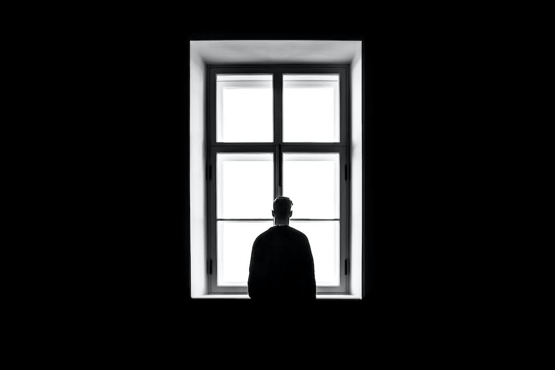 A black-and-white image of a person standing in front of a window feeling winter loneliness and a black foreground.